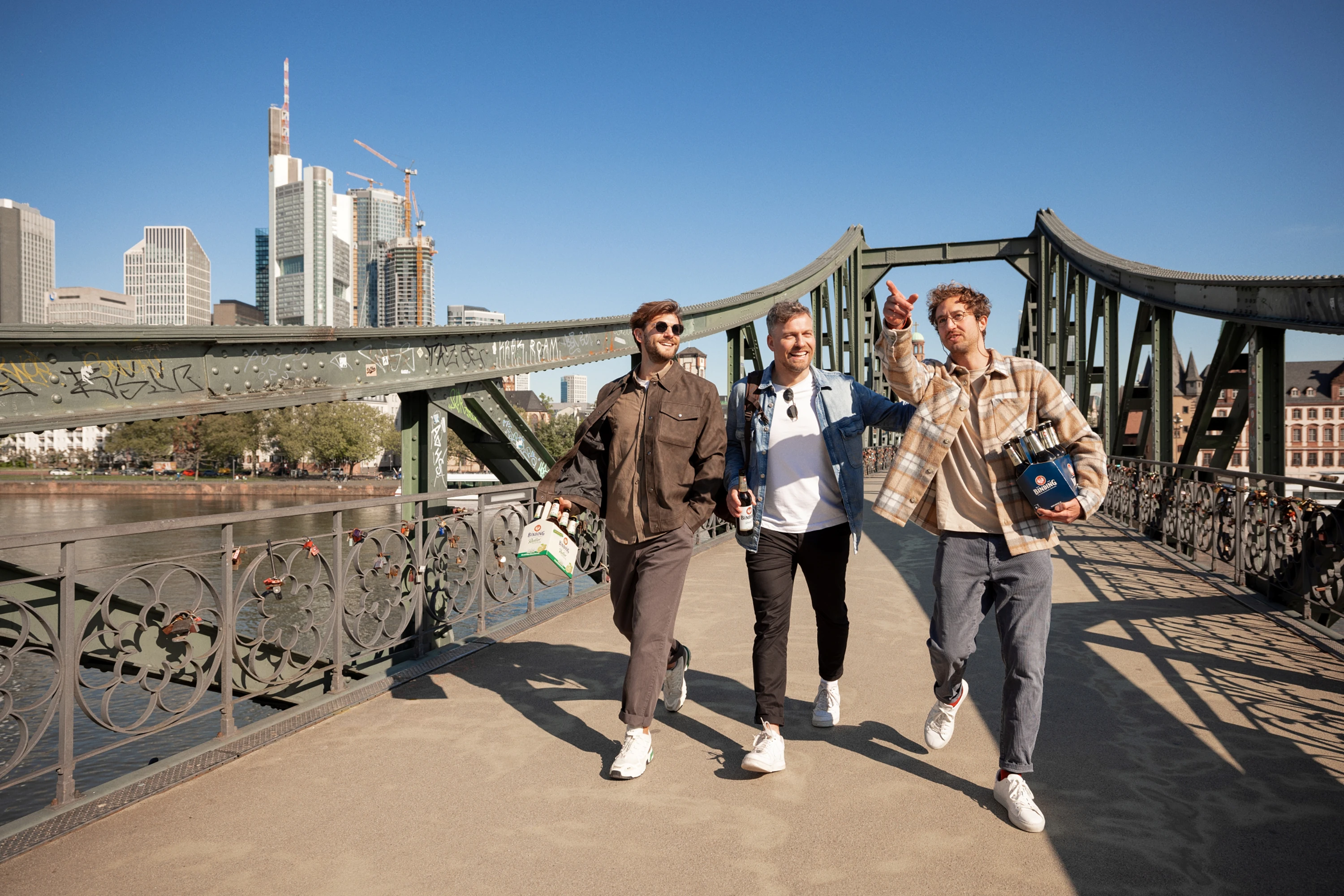 Drei Männer spazieren lächelnd über eine Brücke mit Stadtskyline im Hintergrund.