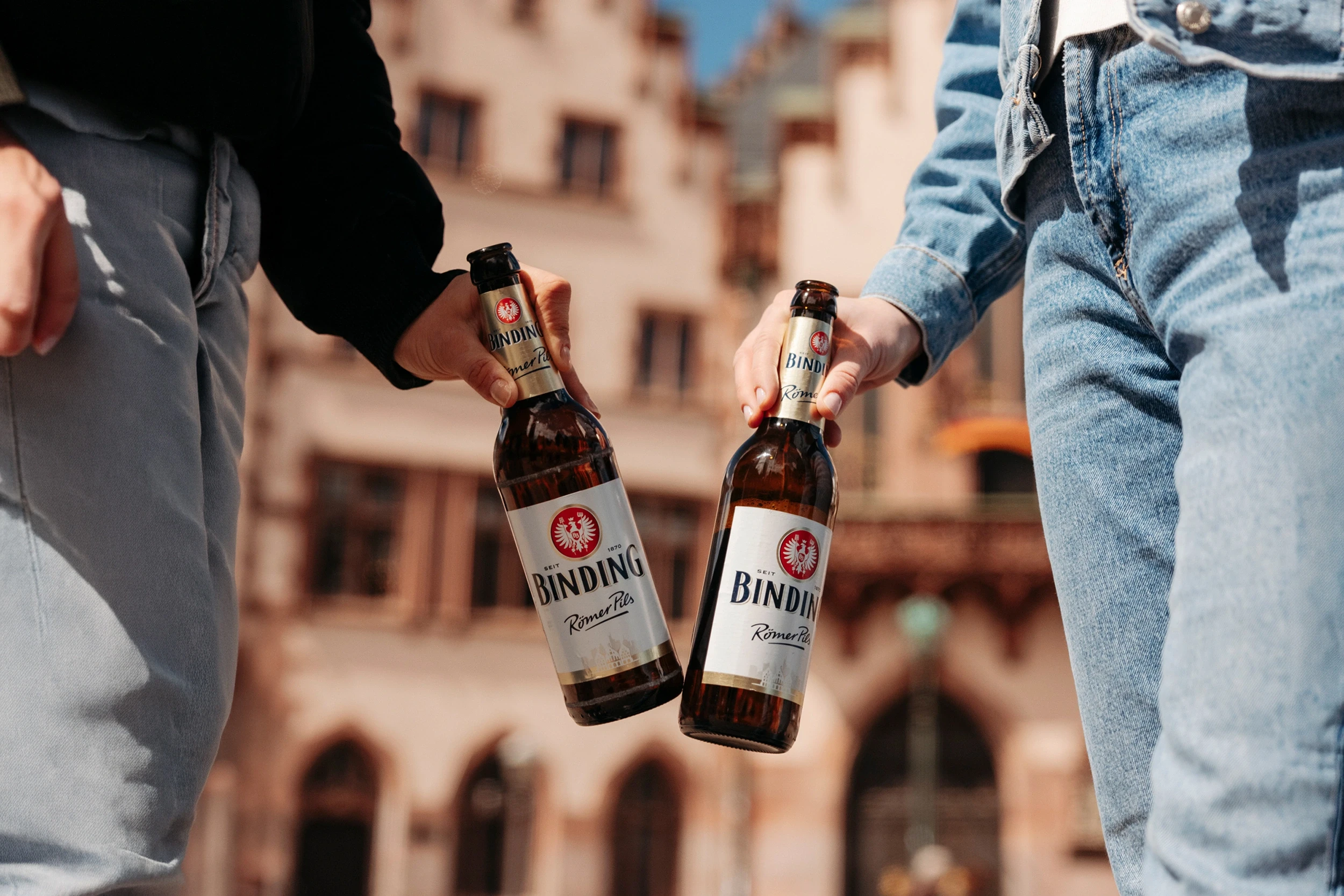 Zwei Personen stoßen mit Flaschen Binding Römer Pils an. Im Hintergrund steht ein historisches Gebäude.