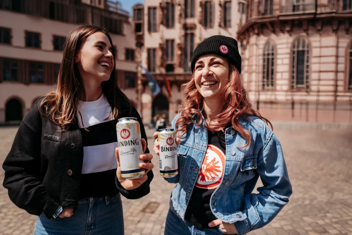 Zwei Frauen mit Dosen Binding Römer Pils stehen auf einem Platz. Historische Gebäude befinden sich im Hintergrund.