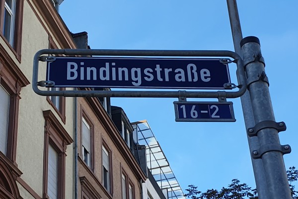 Ein Straßenschild mit der Aufschrift Bindingstraße 16-2 ist an einem Metallpfosten befestigt.