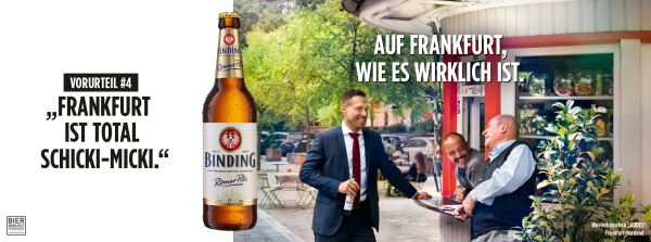 Drei Männer lachen herzlich vor einem Kiosk unter Bäumen.