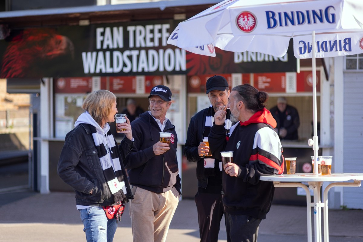 Vier ältere Fußballfans unterhalten sich vor einem Stadion und trinken dabei Bier.