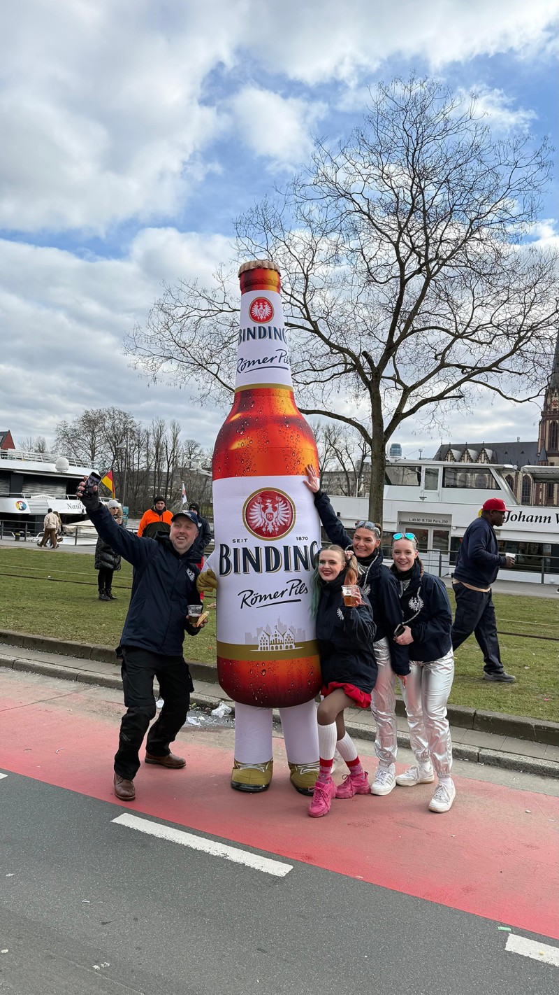 Mehrere Personen stehen lächelnd neben einer Person in einem Kostüm von Binding Römer Pils.