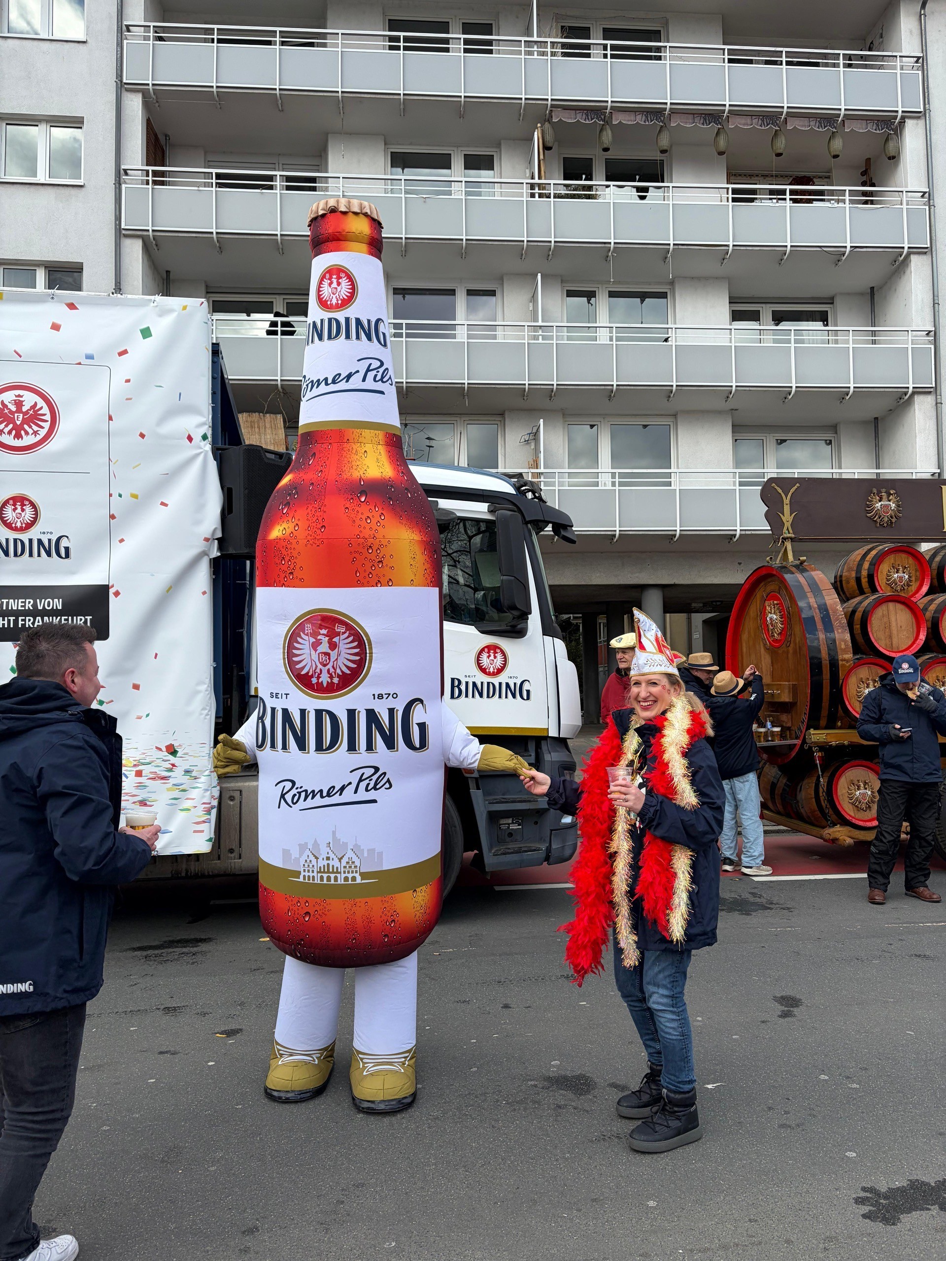 Eine verkleidete Frau steht neben einer Person in einem Kostüm von Binding Römer Pils. Im Hintergrund steht ein Binding-Fastnachtsumzugswagen und ein Wagen mit mehreren Bierfässern.