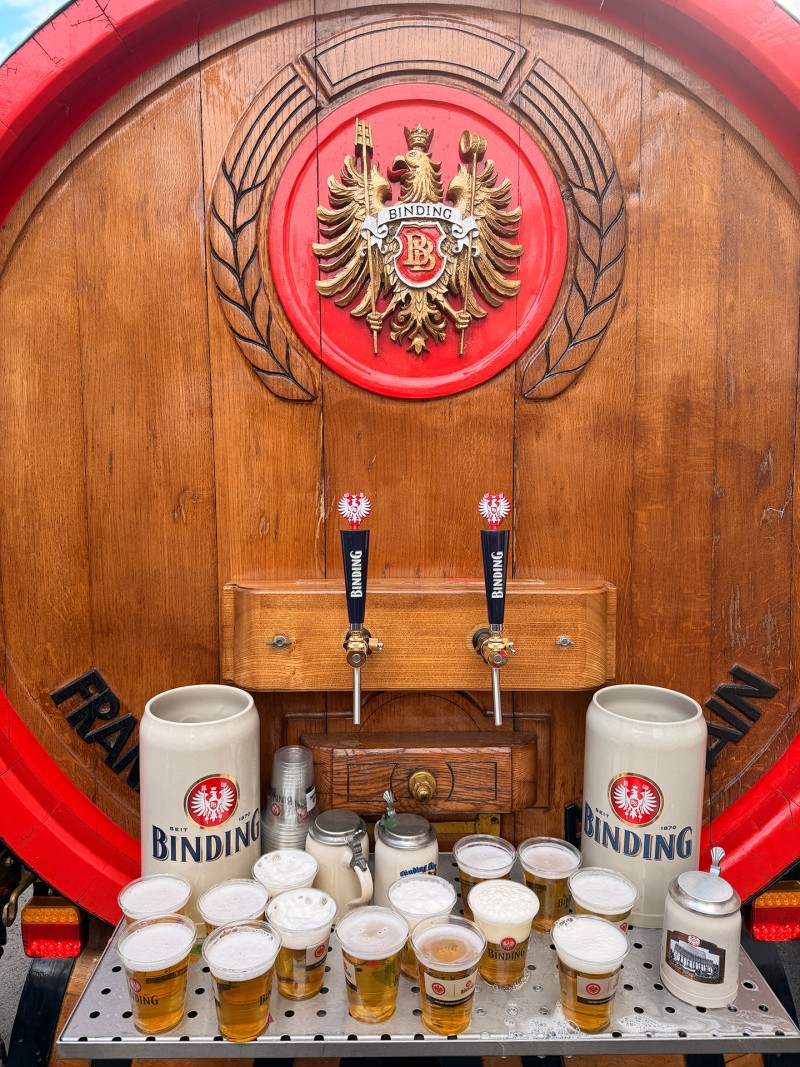 Vor zwei Binding-Zapfhähnen stehen mehrere mit Bier gefüllte Becher und zwei Binding-Bierkrüge.