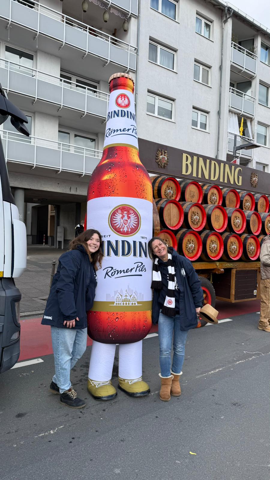 Zwei Frauen stehen neben einer Person in einem Kostüm von Binding Römer Pils. Im Hintergrund steht ein Binding-Fastnachtsumzugswagen und ein Wagen mit mehreren Bierfässern.