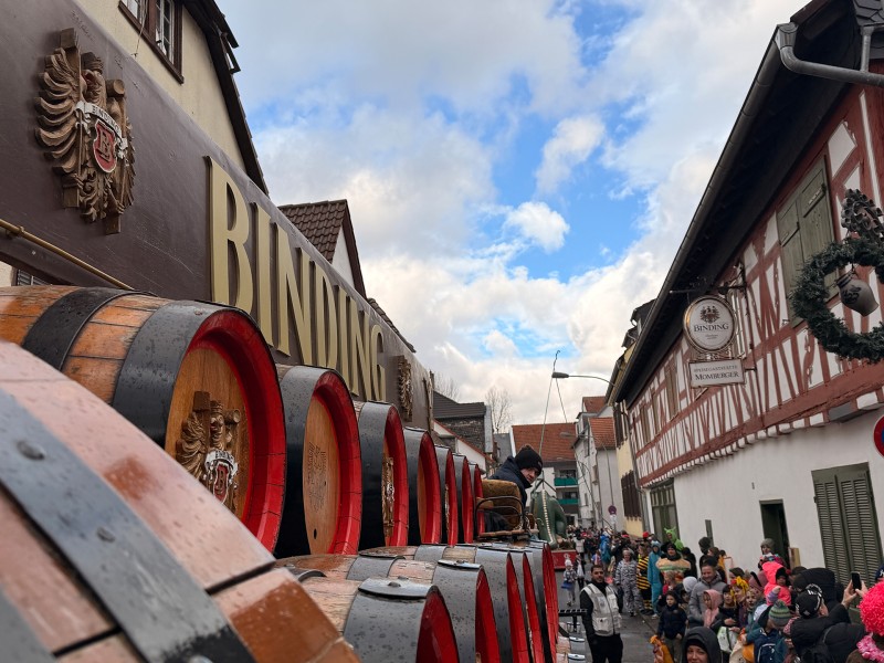 Ein Binding-Fastnachtsumzugswagen mit mehreren Bierfässern darauf fährt an zahlreichen verkleideten Menschen vorbei.