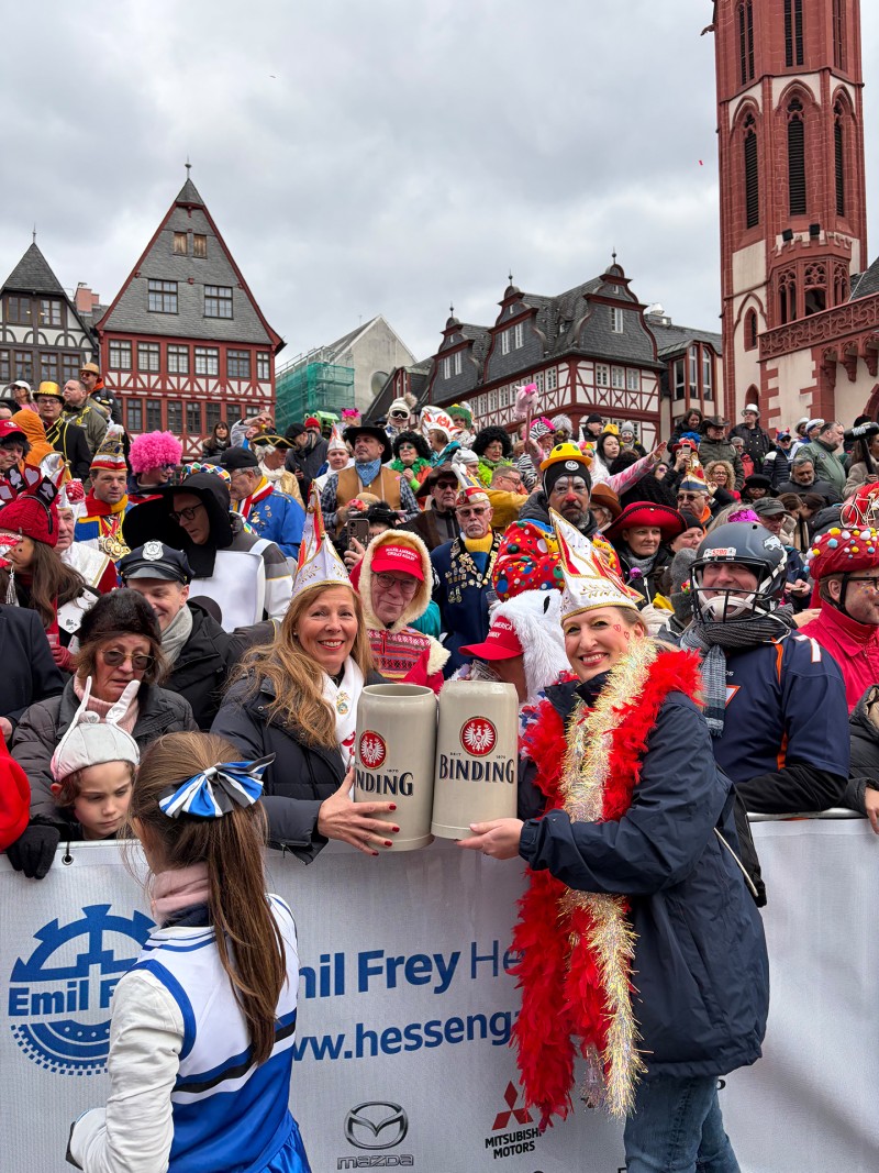 Zwei Frauen stehen vor einer versammelten Menge von verkleideten Fastnachtsbesuchern und halten jeweils einen Binding-Bierkrug in den Händen.