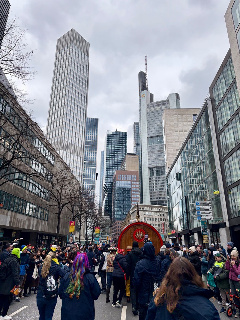 Zahlreiche verkleidete Personen nehmen an einem Fastnachtsumzug in Frankfurt teil. Im Hintergrund sind einige Hochhäuser.