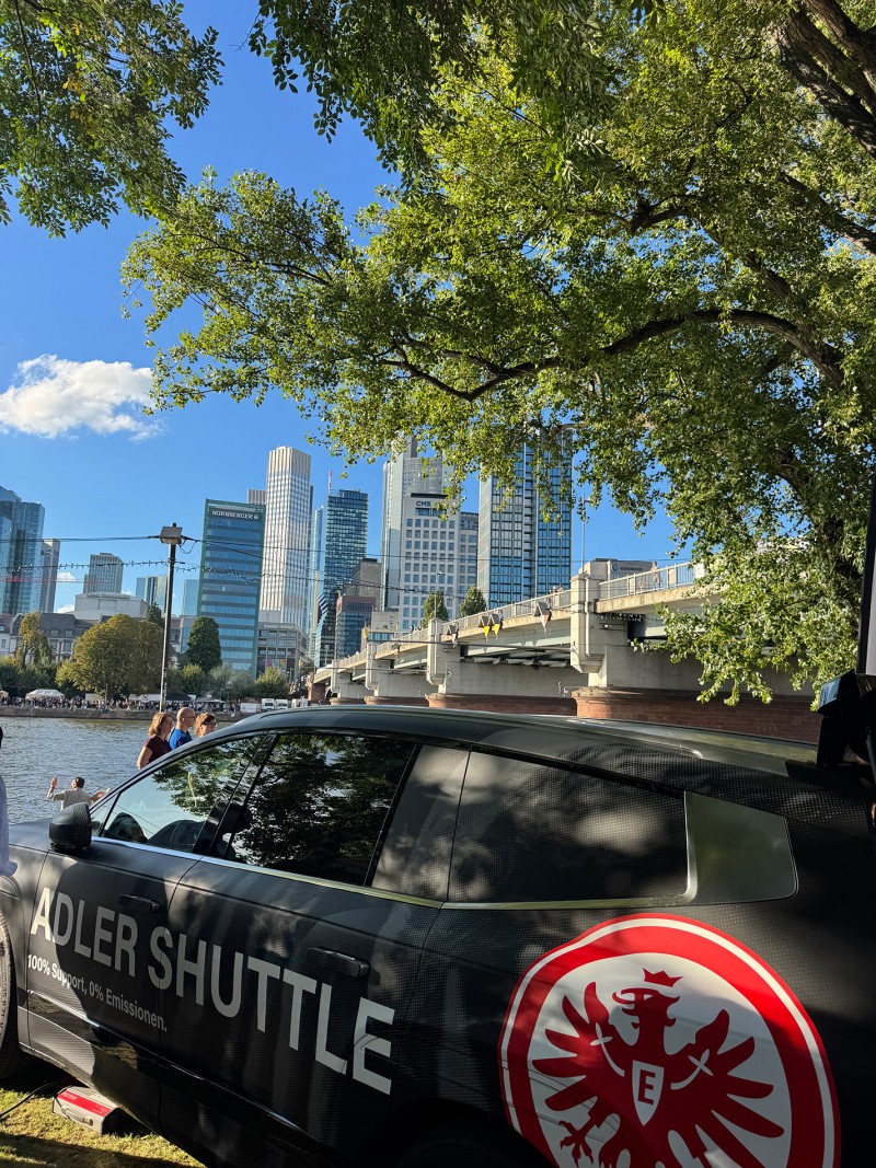 Im Vordergrund steht ein Fahrzeug mit der Aufschrift Adler Shuttle, im Hintergrund die Frankfurter Skyline.