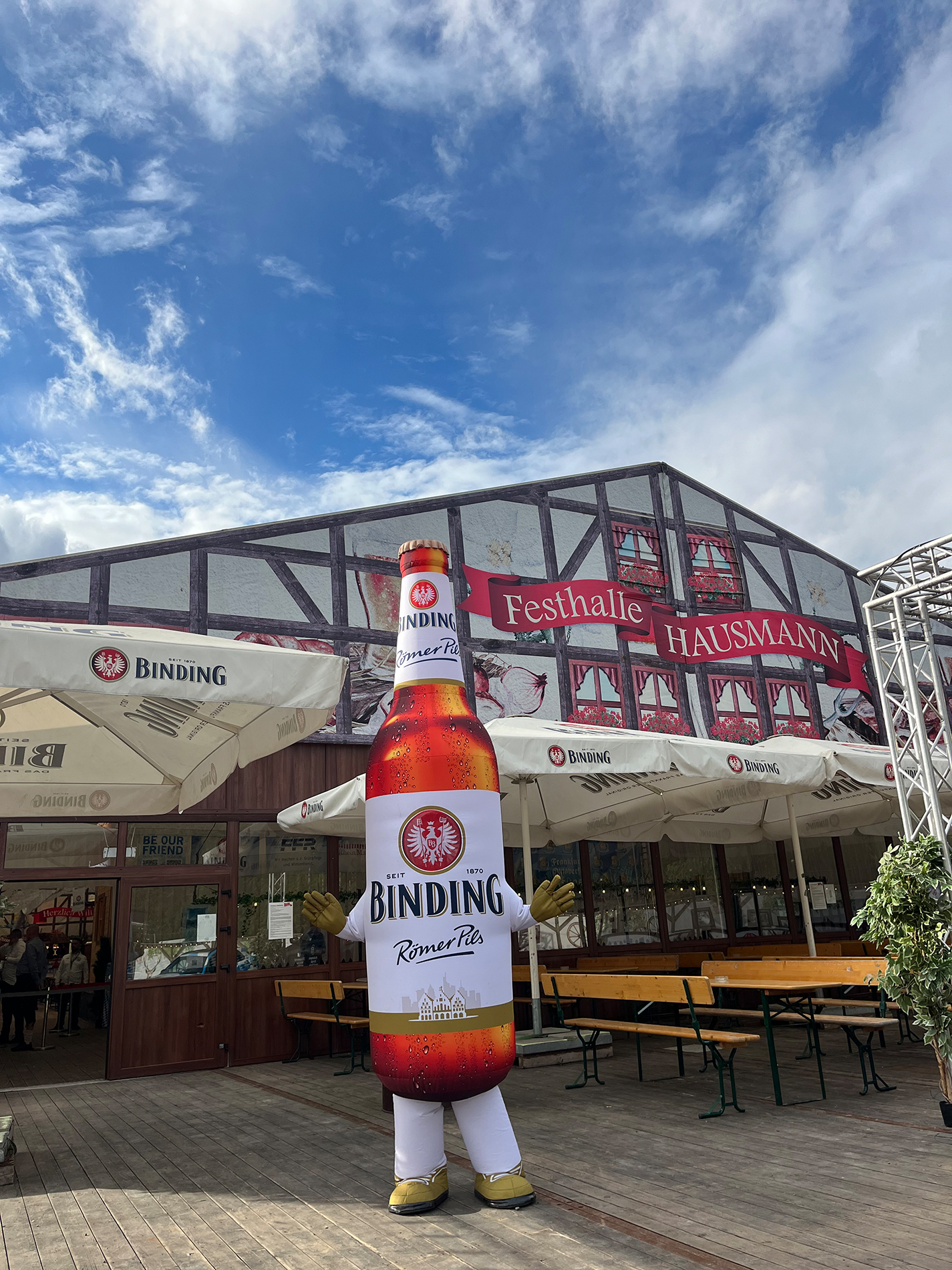 Ein übergroßes Bierflaschen-Maskottchen steht vor einem Bierzelt.