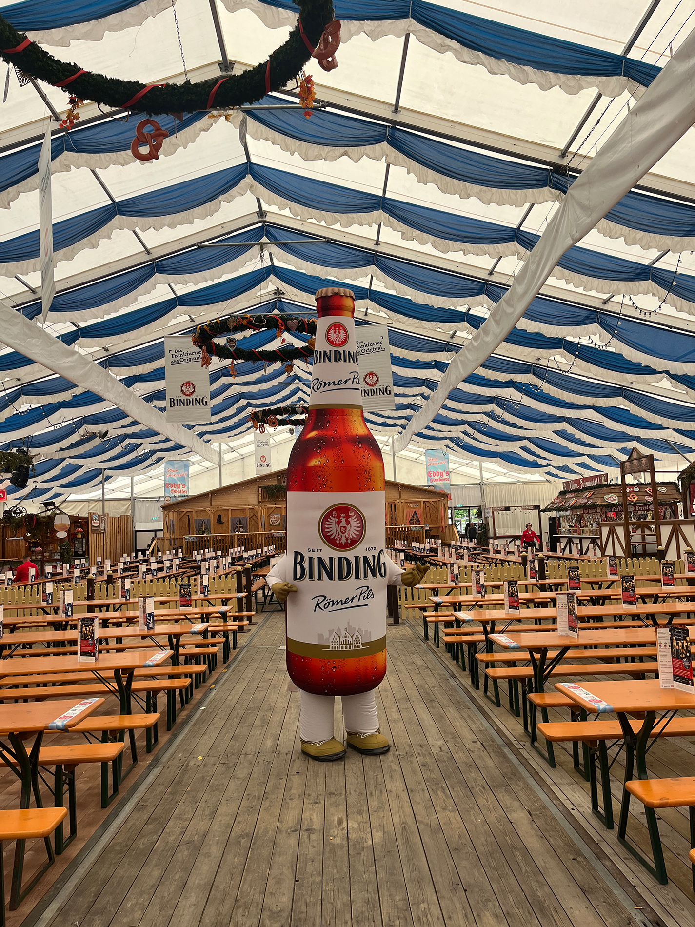 Ein übergroßes Bierflaschenmaskottchen steht in einem Bierzelt.