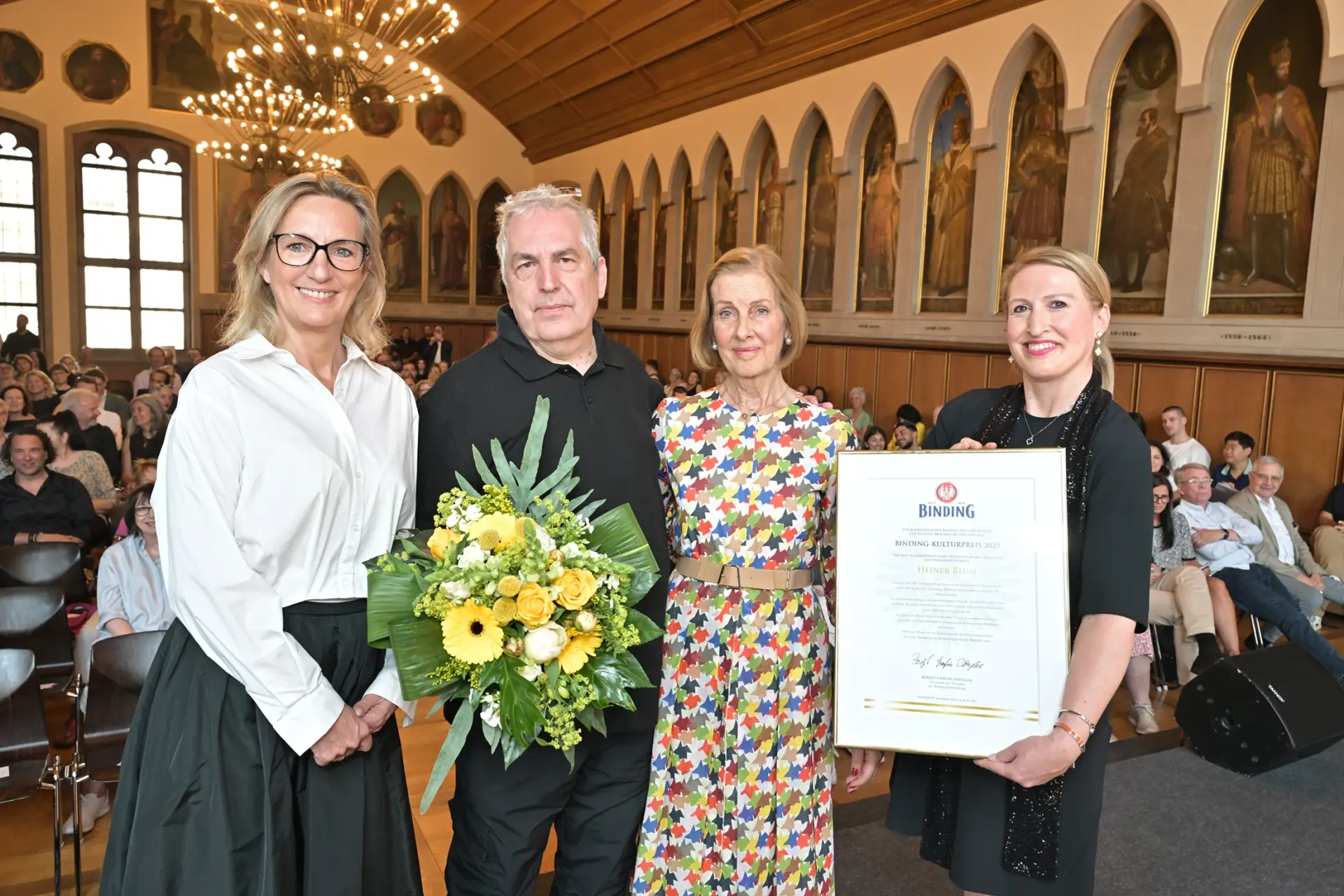 Vier Personen stehen nebeneinander, ein Mann hält einen Blumenstrauß, eine Frau eine Urkunde. Im Hintergrund ein Publikum im Saal.