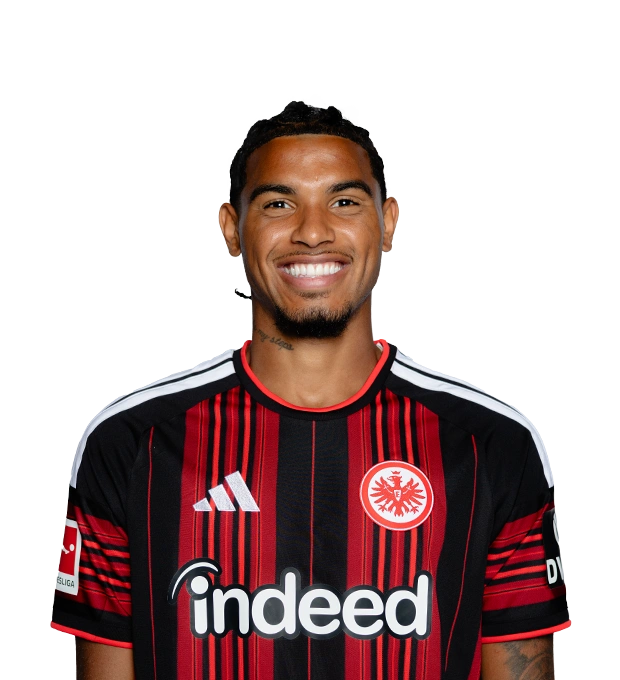Mann mit geflochtenen dunklen Haaren, Kinnbart und breitem Lächeln trägt ein Eintracht Frankfurt Fußballtrikot.