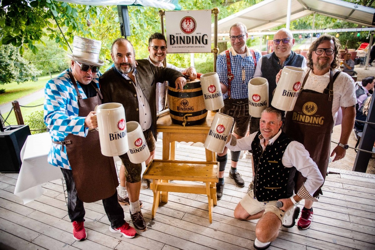 Sieben Männer in traditioneller und lässiger Kleidung halten große Bierkrüge bei einem Fest.