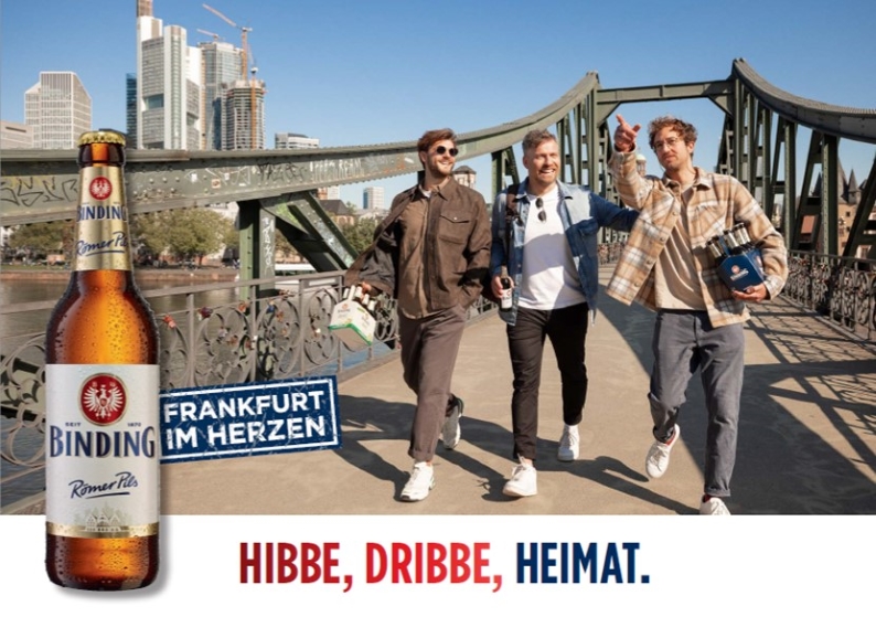 Drei Personen laufen über den eisernen Steg in Frankfurt und trinken dabei Binding Bier.