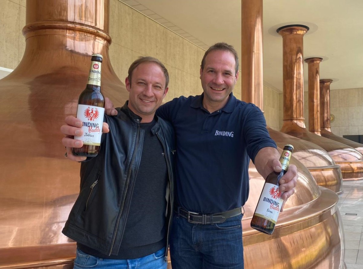 Zwei lächelnde Männer halten Flaschen vor einem großen Kupferkessel. Sie tragen dunkle Kleidung.